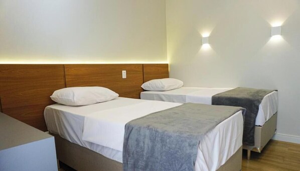 A hotel room with two single beds, each with a white mattress and pillow, and a grey blanket. The room has a wooden headboard and two wall-mounted lights.