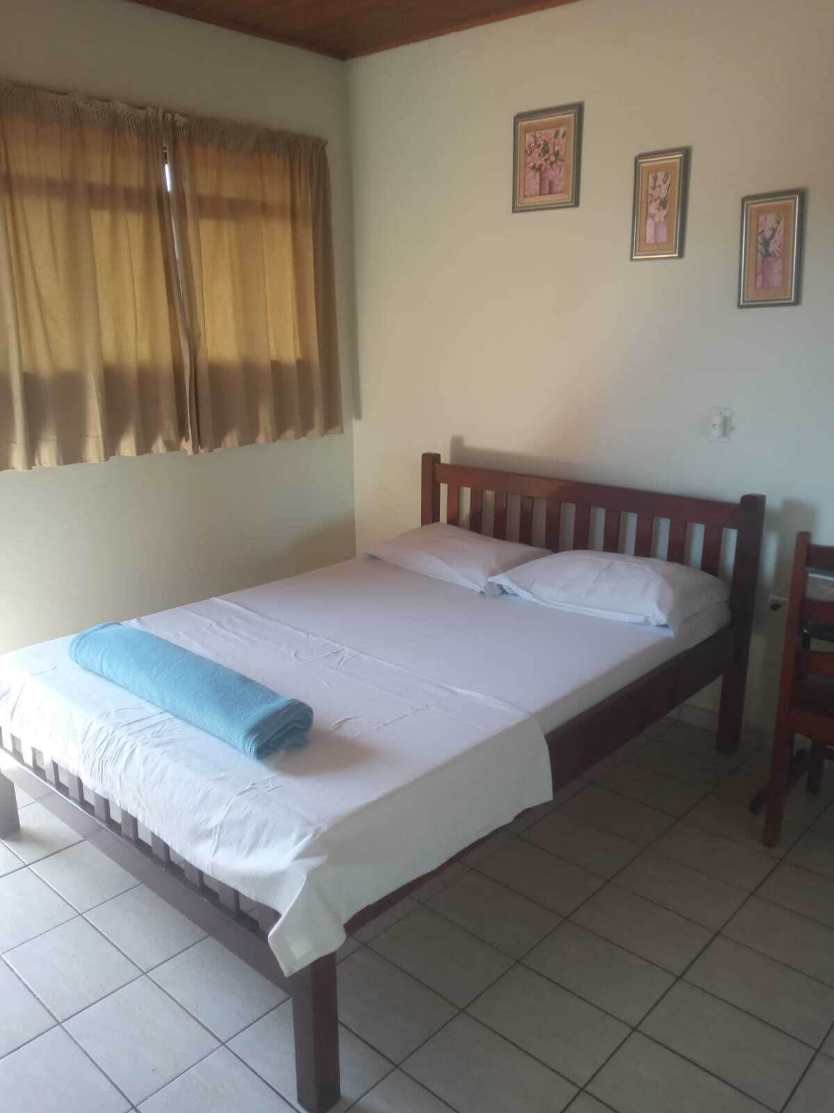 A single bed with a wooden headboard, white bedding, and a blue towel. There are two framed pictures on the wall and a window with curtains.