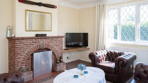 A living room with a brick fireplace, a television on a stand, leather armchairs, and a large window with curtains. - Huge Peaceful 3Bed With FREE Parking (West Sussex)