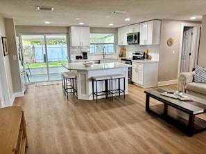 A modern kitchen with white cabinets, a central island with stools, a dining area with a wooden table, and a sliding glass door leading to a backyard. - Fully Renovated Coastal Retreat 3BR w/ Garage & Screened Lanai (Apollo Beach)