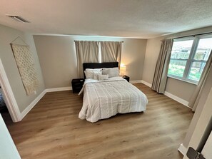 Une chambre à coucher avec un grand lit, une table de chevet, une lampe fixée au mur et une fenêtre avec des rideaux.