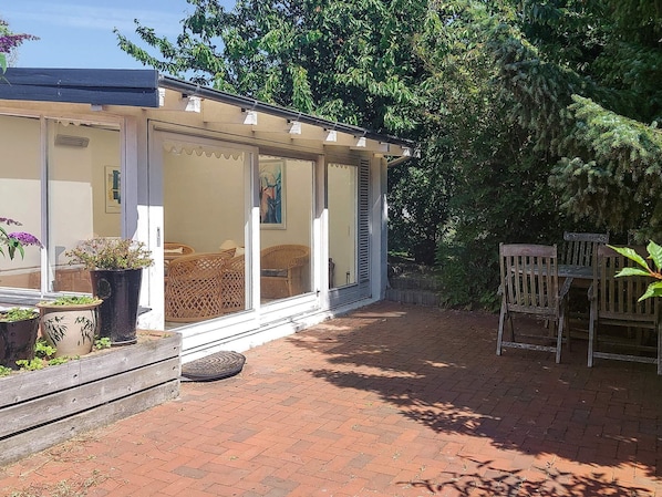 A covered patio area with wicker furniture, potted plants, and a brick pathway leading to a wooden deck.