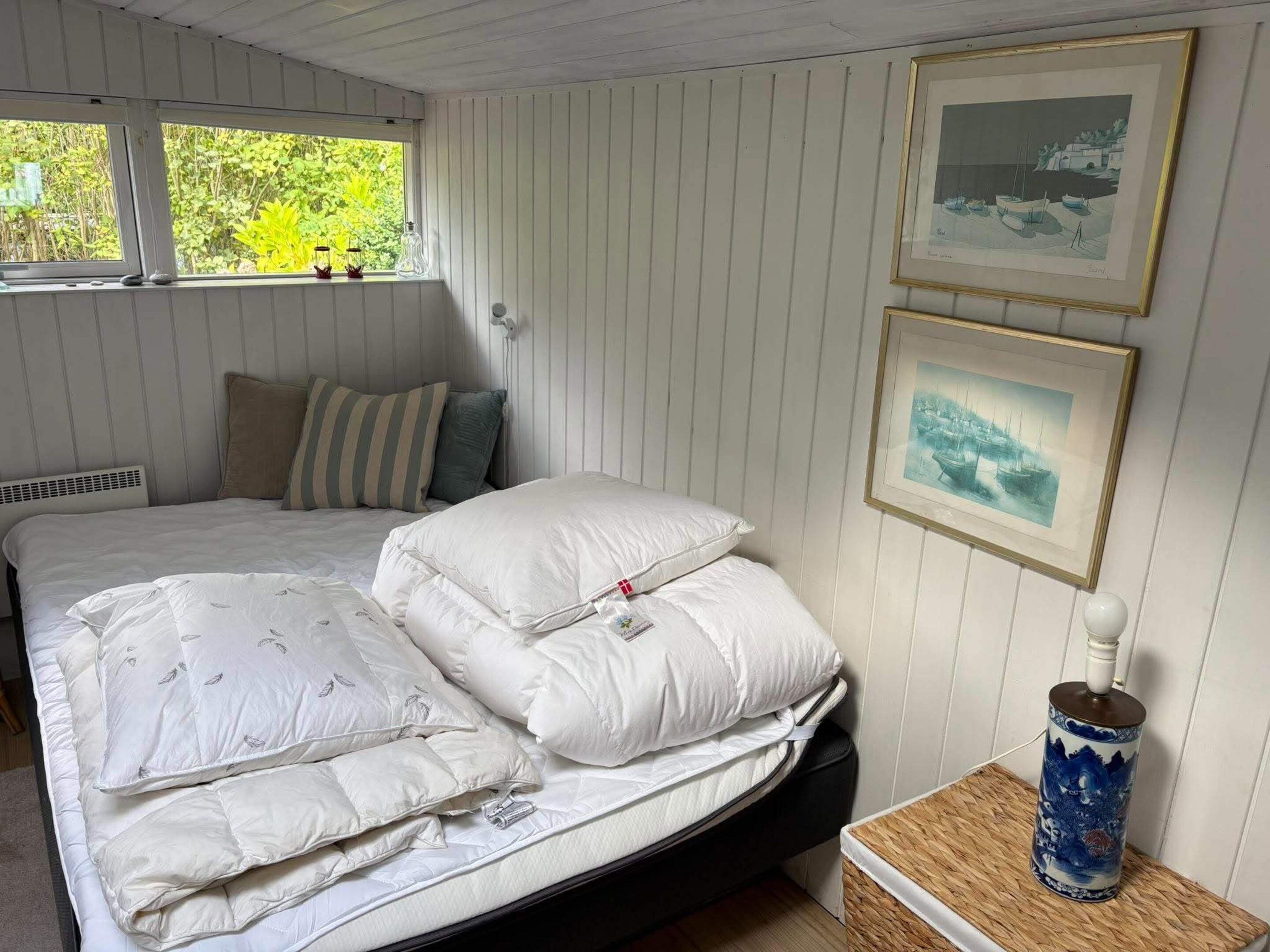 Un dormitorio con cama, almohadas, una mesita de noche con un florero y dos cuadros en la pared.