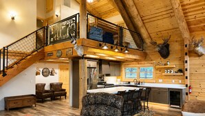 A spacious living area with a high ceiling, a staircase, a kitchen with a large island, and a deer head mounted on the wall.
