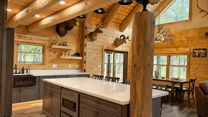 A rustic cabin interior with a large wooden dining table, a kitchen with dark cabinets, and a ceiling with wooden beams and mounted animal heads.