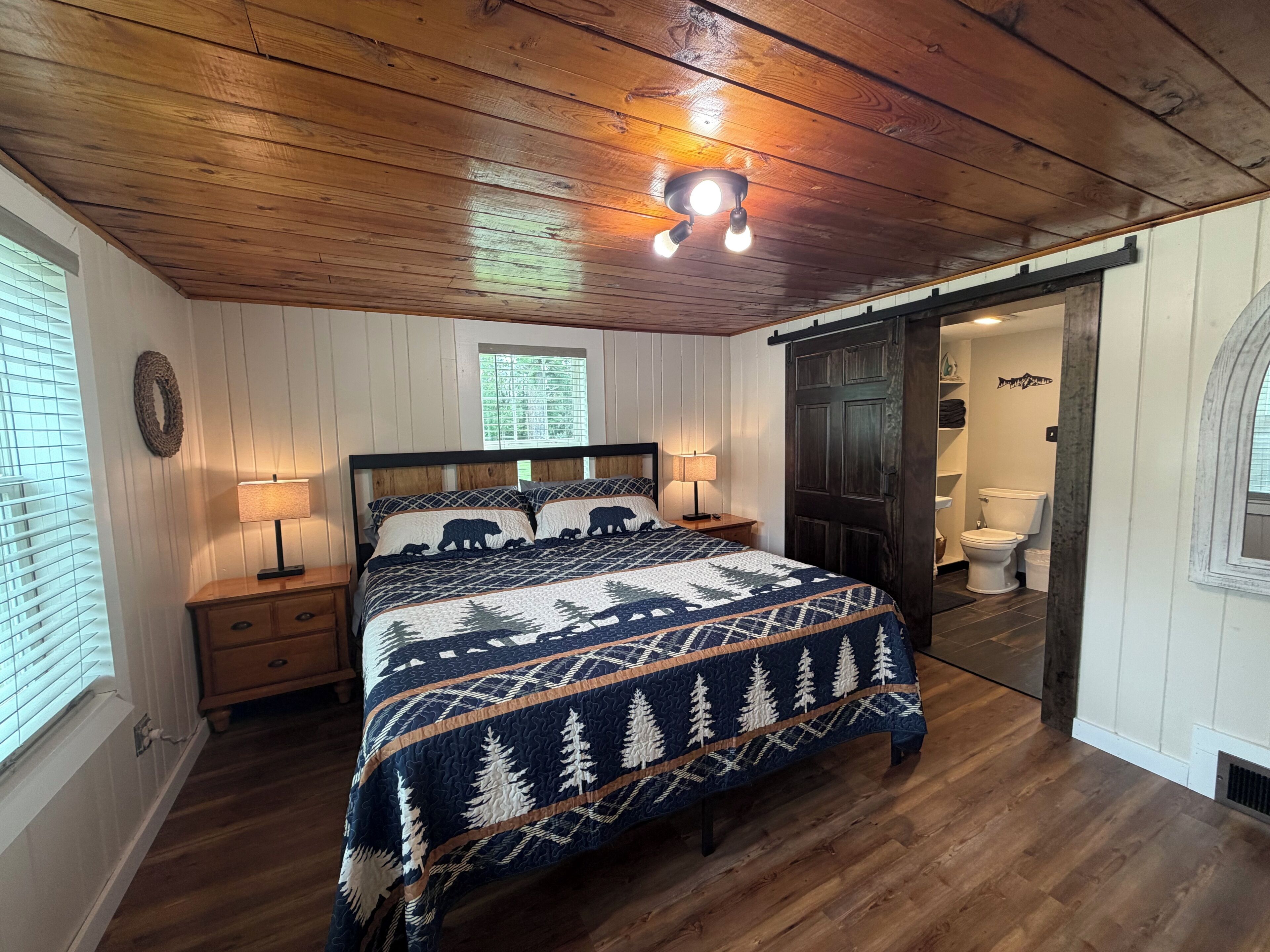 Un dormitorio con techo de madera, una cama con edredón de temática forestal, una mesita de noche con lámpara y vista al baño.