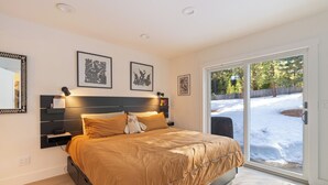 A bedroom with a bed, bedside tables, wall-mounted lamps, framed artwork, and a sliding glass door leading to a snowy outdoor area. - Mountain Modern Retreat with Saltwater Hot tub and Cedar Sauna (Truckee)