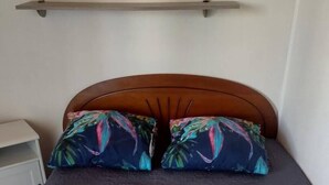 Een bed met een bedovertrek met een patroon en twee kussens met een tropisch ontwerp, een houten hoofdbord, en een nachtkastje aan de linkerkant.