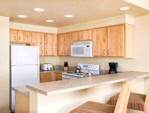 A kitchen with wooden cabinets, a white refrigerator, a microwave, a stove, and a coffee maker. - Seaside!! Condo with view, Balcony, Grill, just steps to the beach! (Seaside)
