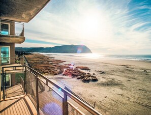 Vista a la playa desde un balcón, con cielo despejado, arena dorada y montañas a lo lejos.