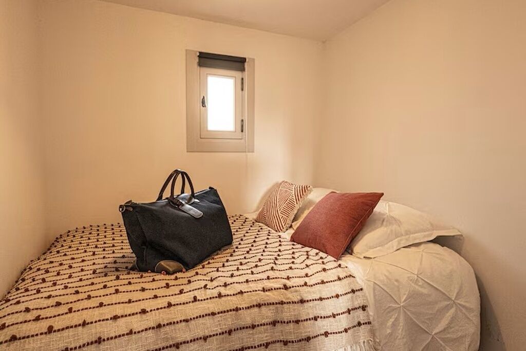 Ein Einzelbett mit einem gemusterten Bettbezug, eine dunkel gefärbte Tasche auf dem Bett und ein Fenster darüber.