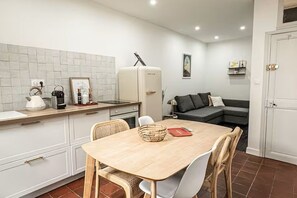 A compact kitchen with a dining table, chairs, a refrigerator, and a sofa in the background.