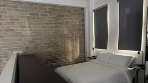 Quarto com parede de tijolos, cama com roupa de cama branca, escrivaninha e duas luminárias de cabeceira.