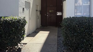 Um beco estreito que leva à entrada de um edifício, com um portão de metal, um pequeno arbusto e um caminho pavimentado.