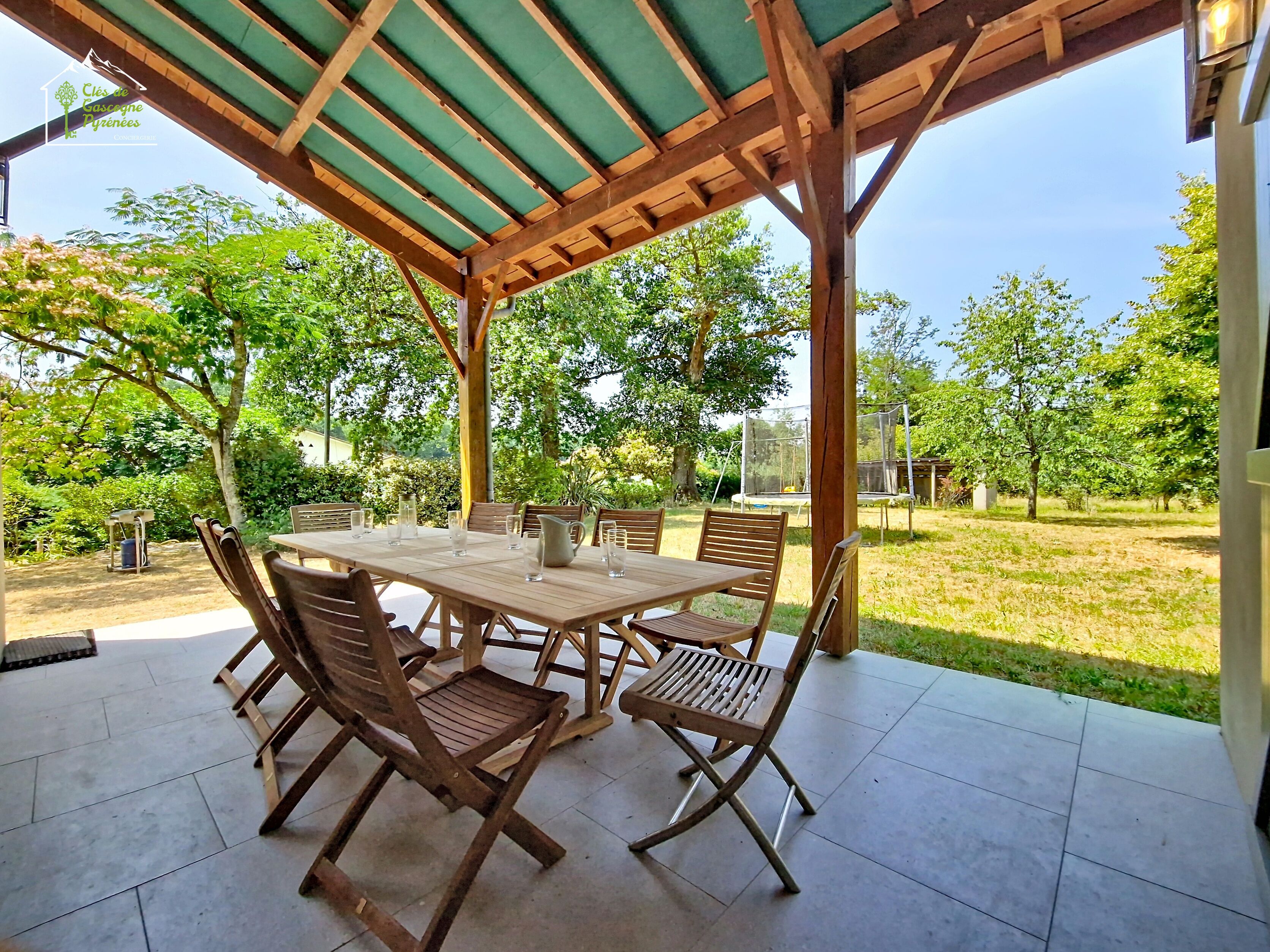 Une terrasse extérieure avec une table et des chaises en bois, un espace couvert doté d’un toit végétalisé et une vue sur une zone herbeuse avec des arbres.