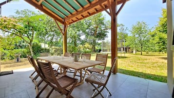 Une terrasse extérieure avec une table et des chaises en bois, un espace couvert doté d’un toit végétalisé et une vue sur une zone herbeuse avec des arbres.