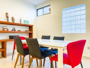 A dining area with a white table, red and blue chairs, a wooden bookshelf with decorative items, and a window with a grid pattern. - Sueños del Mar 2 (Playa del Carmen)