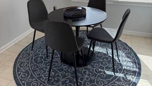 Un comedor con una mesa redonda, cuatro sillas, un tapete estampado, una lámpara colgante y un ventanal con persianas.
