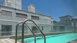 Ein Schwimmbecken mit Metallleiter, einer gefliesten Terrasse und einem Gebäude mit Balkon im Hintergrund.