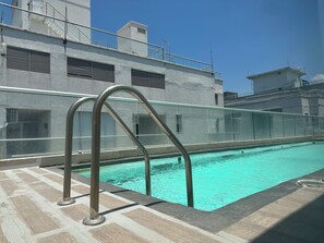 A swimming pool with a metal ladder, a tiled deck, and a building with a balcony in the background. - Edificio Sagitario - Calidad & Precio (Cartagena de Indias)