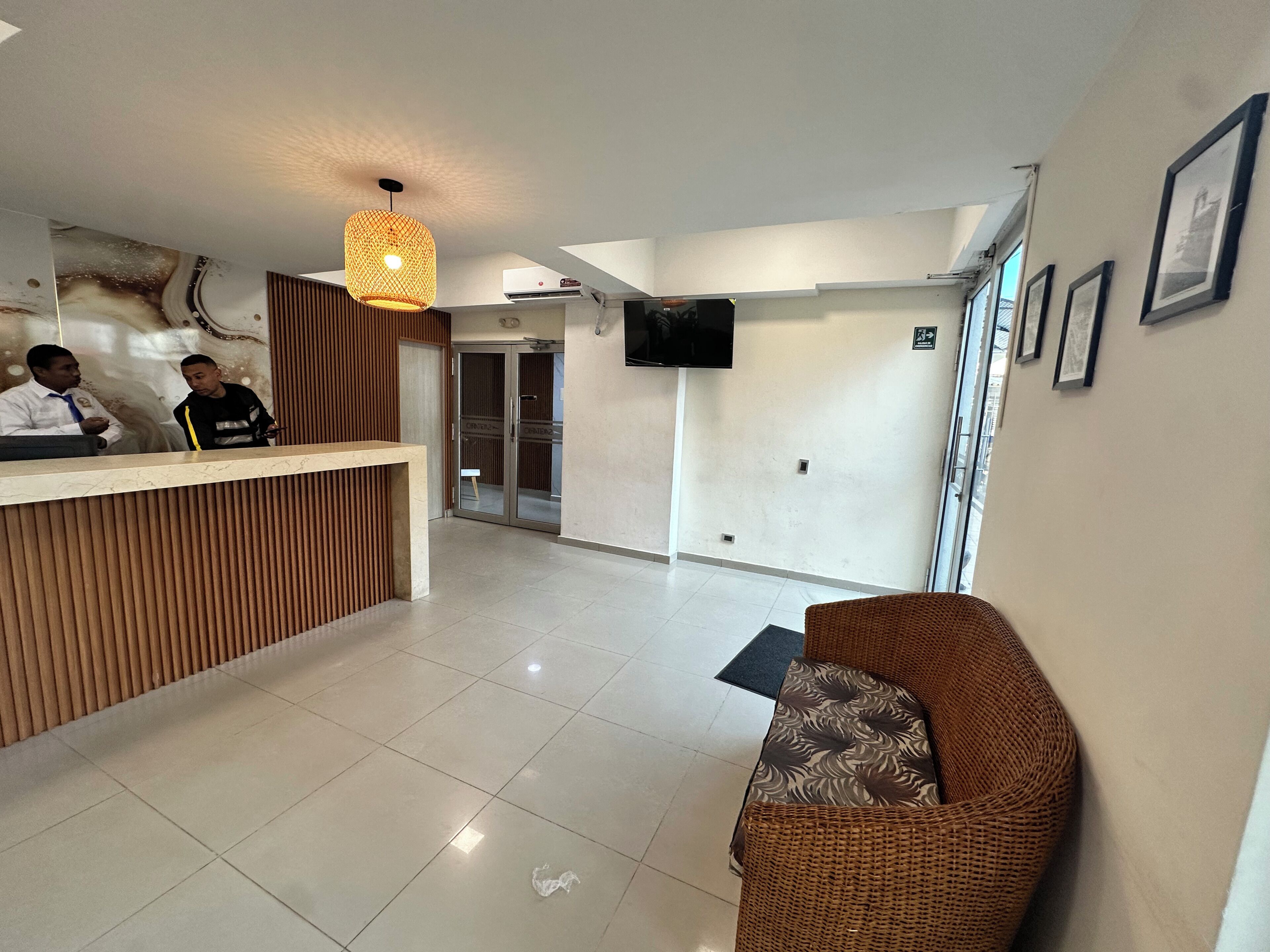 A reception area with a wooden reception desk, a wicker sofa, a flat-screen TV mounted on the wall, and a tiled floor.