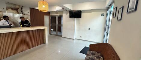 A reception area with a wooden reception desk, a wicker sofa, a flat-screen TV mounted on the wall, and a tiled floor.