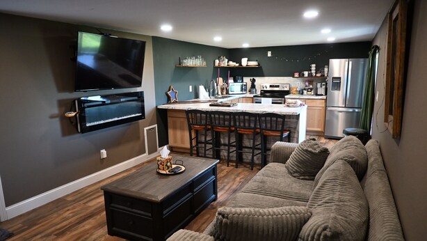 A modern living room with a flat-screen TV, a wooden coffee table, a gray sofa, and a kitchen area with a refrigerator, microwave, and bar stools.