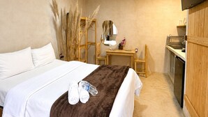 Quarto de hotel com cama, escrivaninha, espelho, televisão e cozinha compacta.