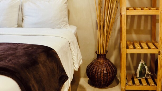 Cama com roupa de cama branca, manta de tecido escuro, um vaso de vime e uma prateleira de madeira com um ferro pequeno.