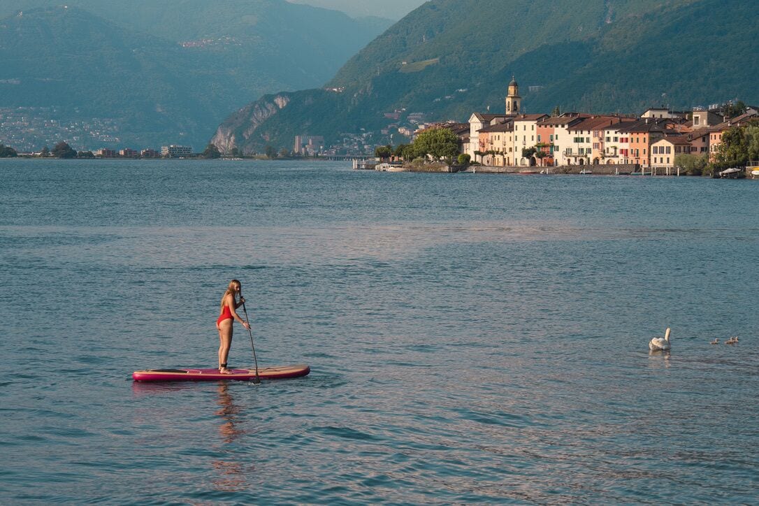 A person paddleboarding on a calm lake with a town and mountains in the background.