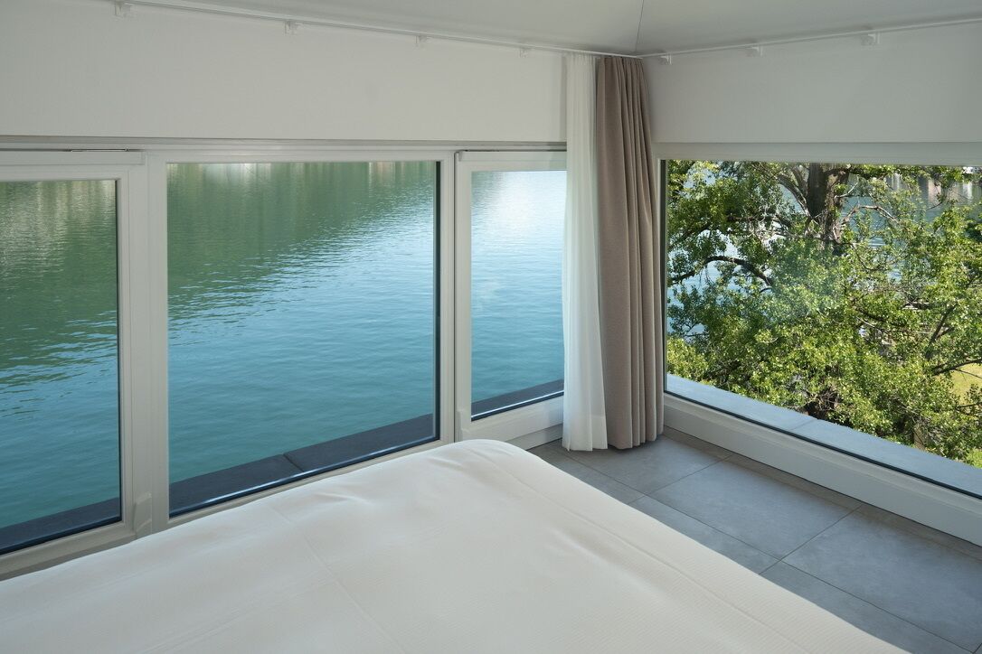 A bedroom with a large window overlooking a body of water and greenery.