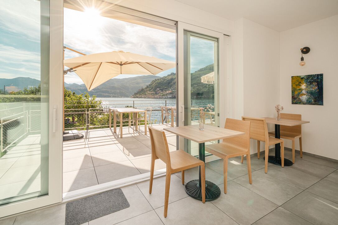 Una camera moderna con un balcone che si affaccia su un lago, arredata con tavoli e sedie in legno e una grande finestra che offre una vista panoramica.