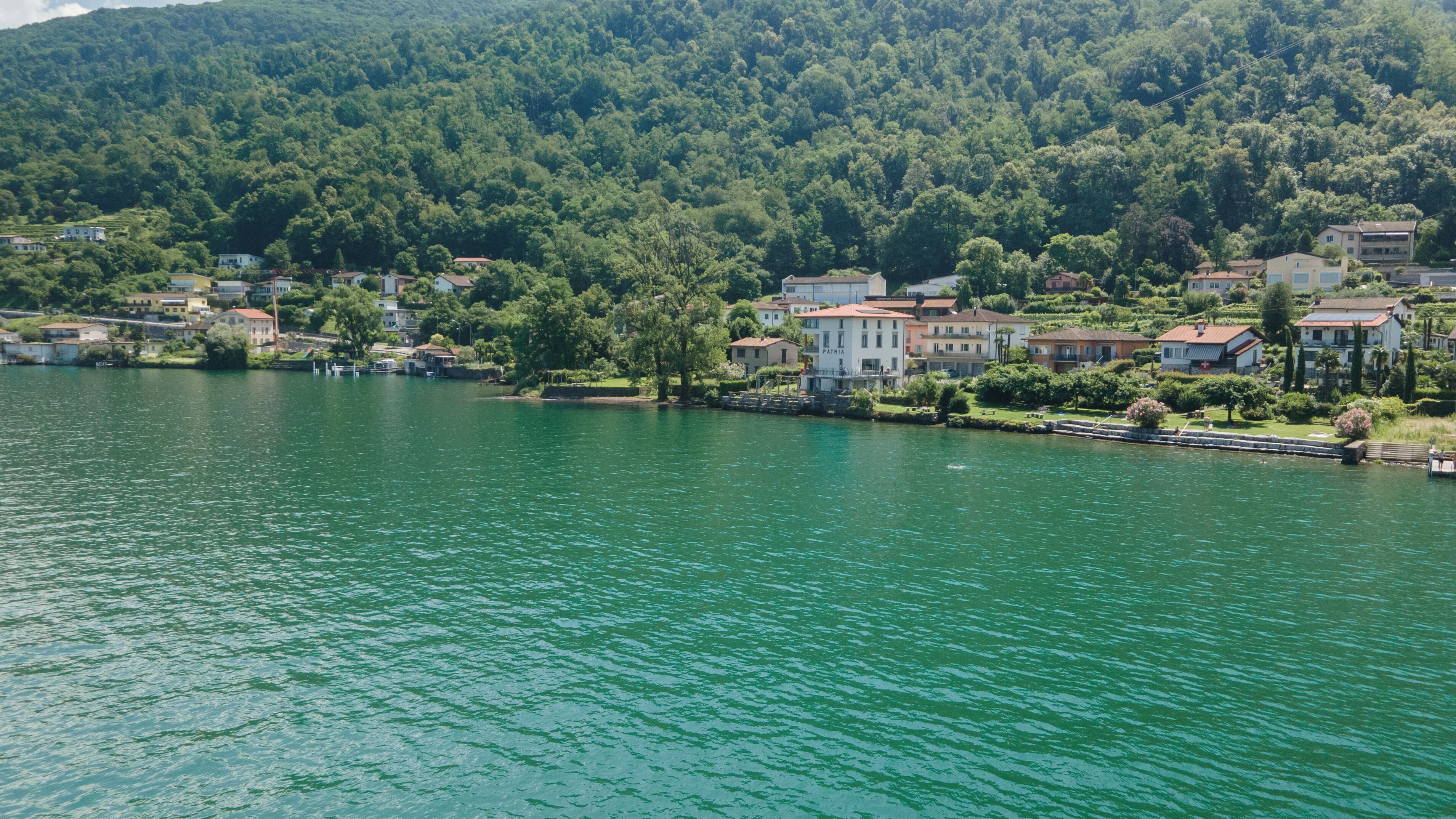 Une ville au bord d’un lac, avec des bâtiments qui bordent la rive, un quai et une colline boisée en arrière-plan.