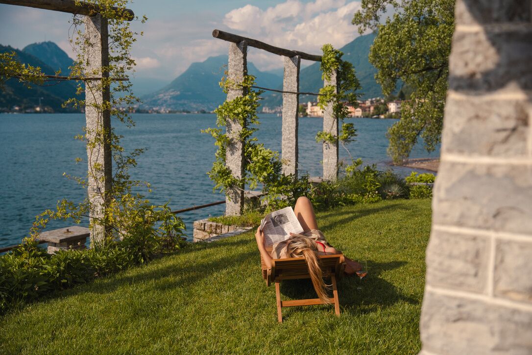 A person relaxing on a wooden chair by a lakeside with stone pillars and a view of mountains.