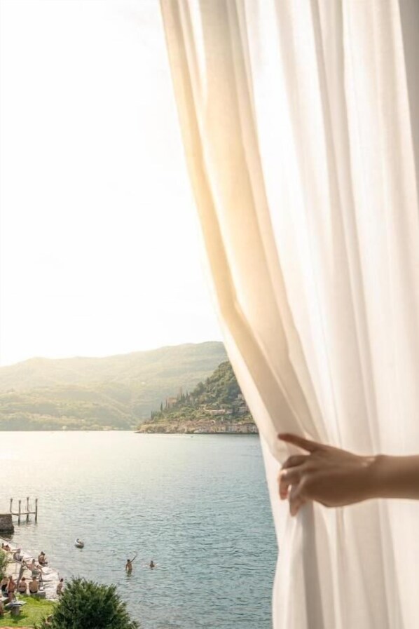 A hand partially opening a curtain to reveal a lake view with boats and people.