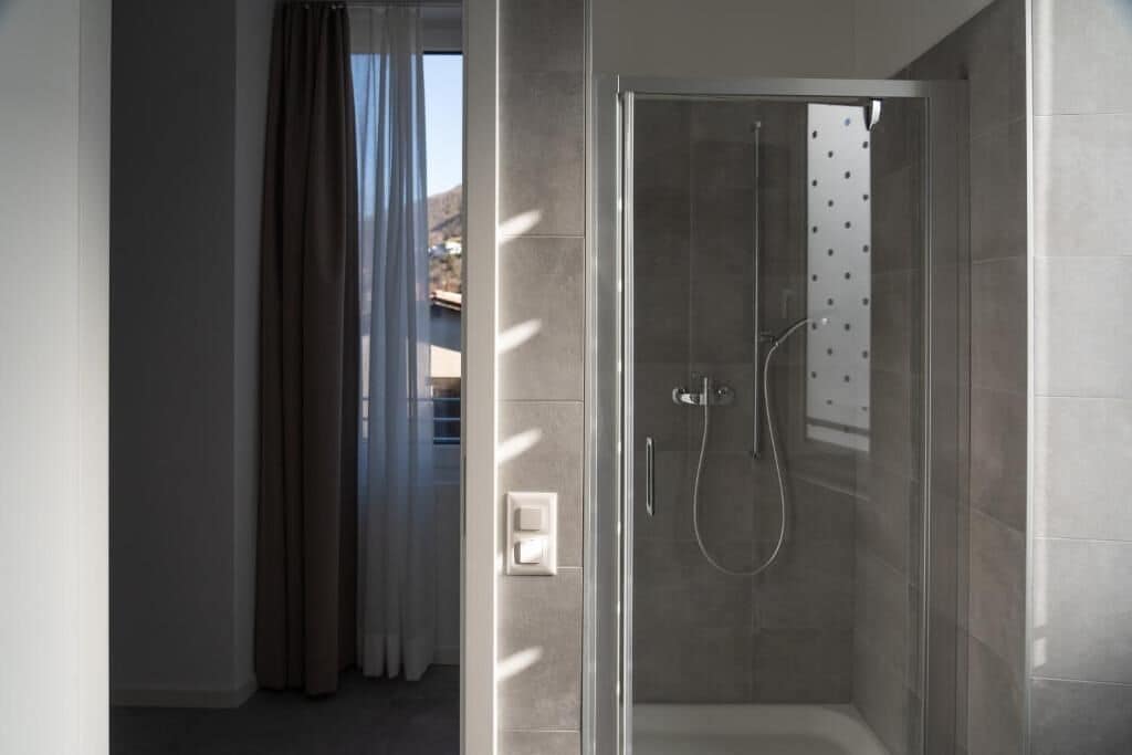 A modern bathroom with a glass-enclosed shower, a visible light switch, and a view of a building outside.