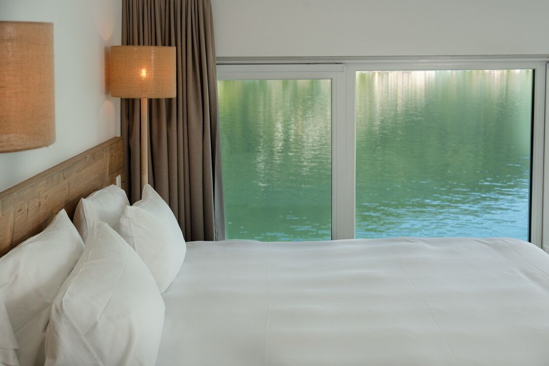 A hotel room with a large bed, a nightstand, a lamp, and a view of water through a window.