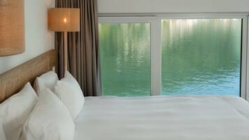 A hotel room with a large bed, a nightstand, a lamp, and a view of water through a window.