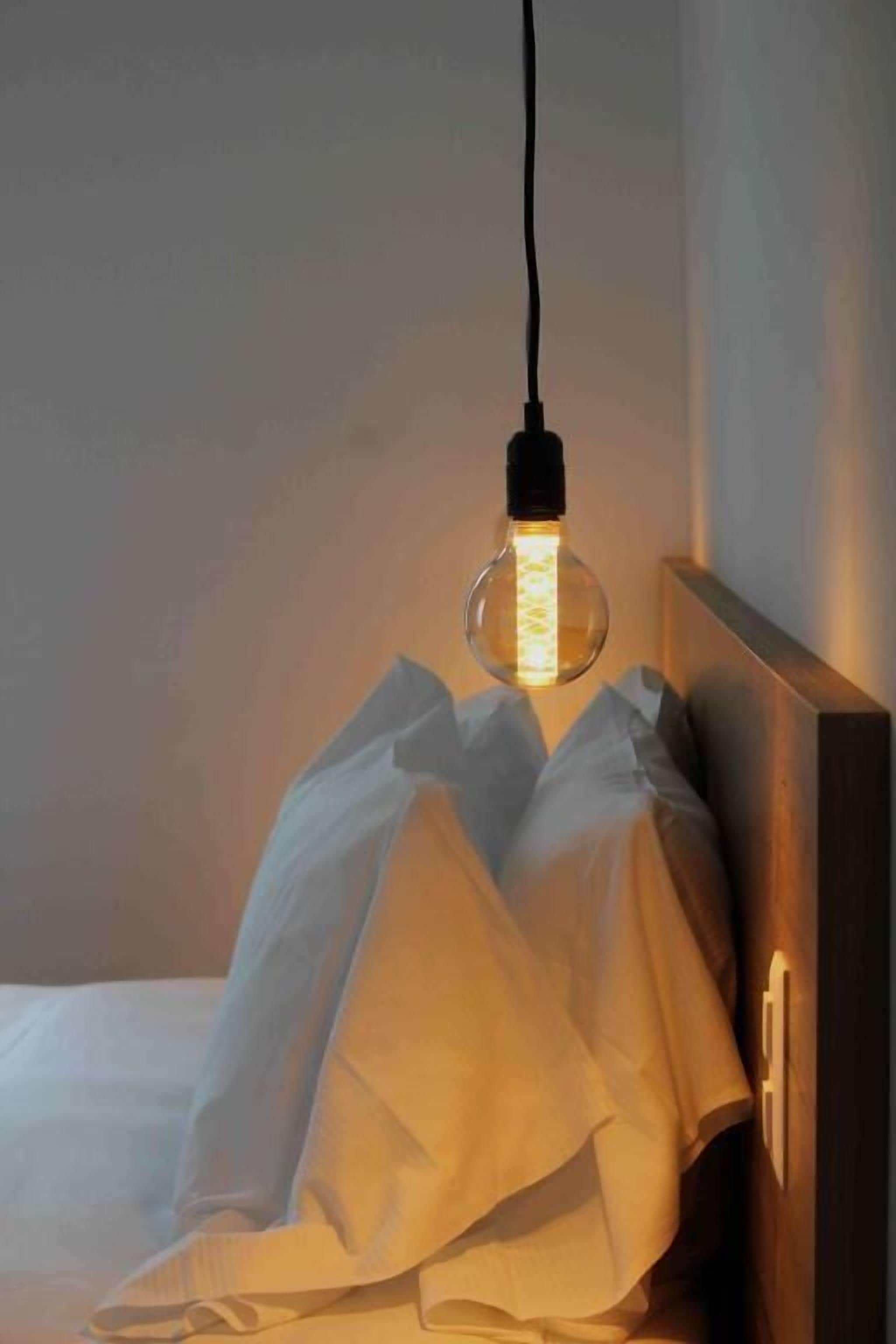 Un lit avec un drap blanc et une lampe dotée d’un abat-jour en verre transparent, suspendue au-dessus.