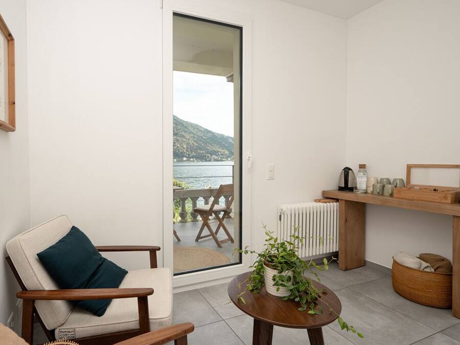 Un espacio habitable moderno con vista a un cuerpo de agua, una silla de madera, una mesita con una planta y un estante con objetos decorativos.