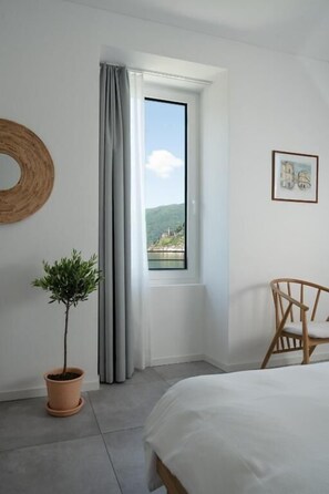 A bedroom with a bed, a window with curtains, a potted plant, a wooden chair, and a wall-mounted circular decoration.
