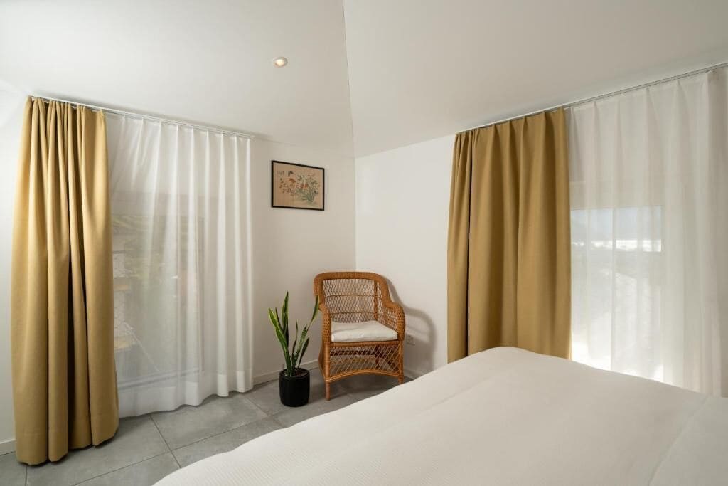 Ein Hotelzimmer mit Bett, Stuhl, Topfpflanze und durchsichtigen Vorhängen.