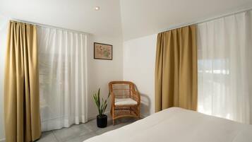 Ein Hotelzimmer mit Bett, Stuhl, Topfpflanze und durchsichtigen Vorhängen.