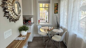 A compact kitchen with a dining area, a white wall with a decorative mirror, a window with a view of a white fence, and a potted plant. - Cozy Ocean Gateway at Spirit Bay Pet-friendly (Sooke)