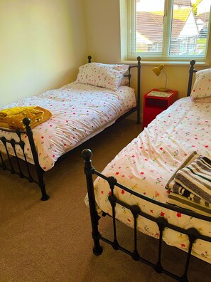 A bedroom with two single beds, a red nightstand, a window with blinds, and a view of a building outside. - Newly renovated house in Polperro with parking and sea views. (Polperro)