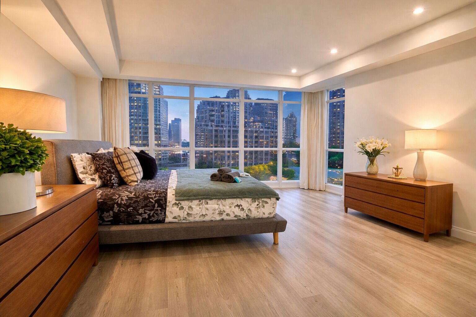 Een moderne slaapkamer met een groot bed, een nachtkastje, een dressoir en uitzicht op de stad door grote ramen.