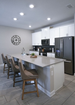 A modern kitchen with white cabinets, a central island with bar stools, a stainless steel refrigerator, and a wall clock. - Luxury Disney Retreat: Private Pool + ChampionsGate Resort Access (Davenport)