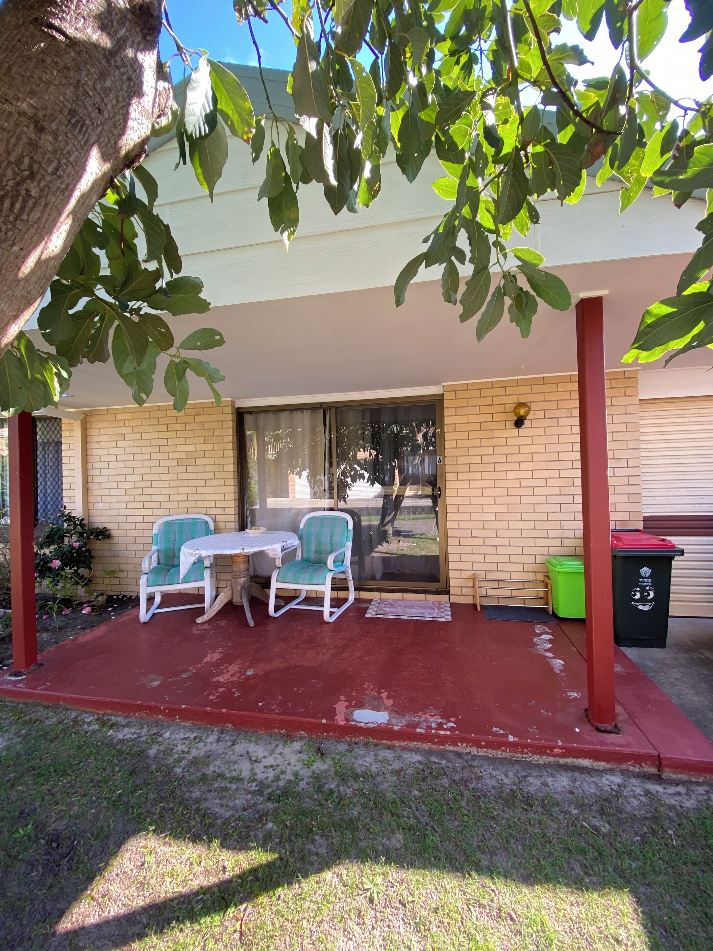 A covered patio with two chairs and a table, a brick wall, a door, and a trash bin.