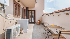Un estrecho pasillo exterior con una mesa, sillas, un refrigerador y un calentador de pared.