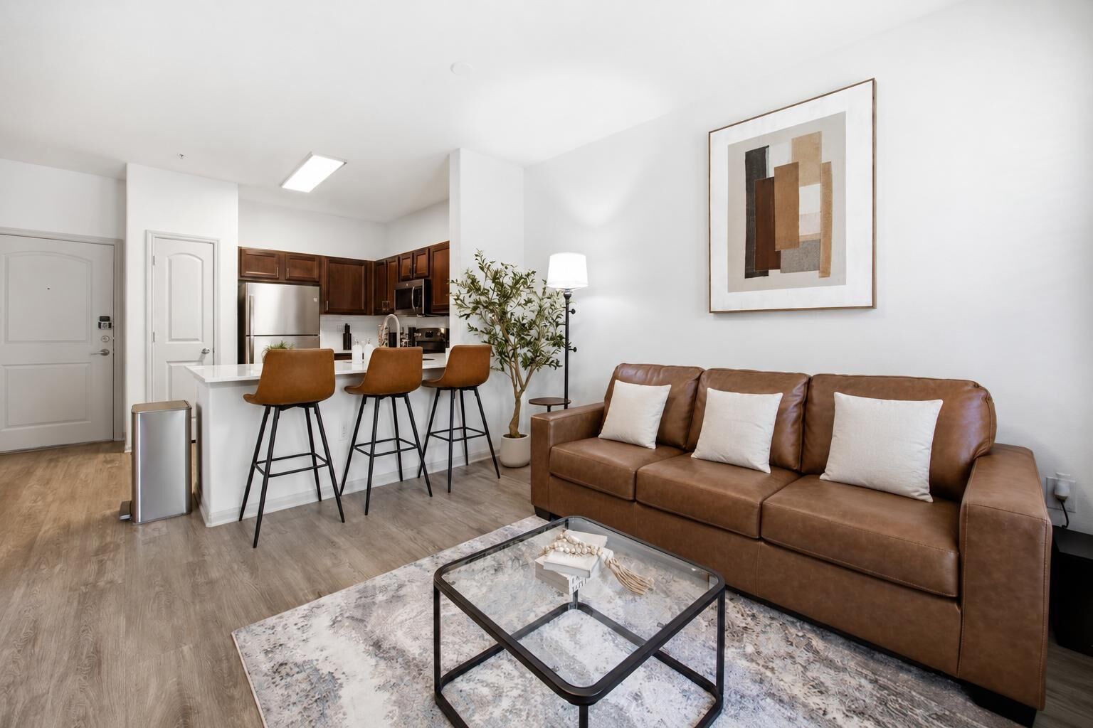 Una sala moderna con un sofá de cuero marrón, una mesa de centro de vidrio y una cocina con armarios de madera y encimera blanca.